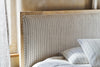 A close-up of the Sanja Stripe Bed - Natural by nkuku, featuring an upholstered, tall cotton headboard with vertical gray stripes and a solid mango wood frame. The bed is dressed in white striped bedding and pillows, with draped curtains at the side.