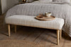 The nkuku Sanja Stripe Upholstered Bench - Grey with natural cotton fabric and wooden legs sits at the beds end. A round wooden tray holds a glass of water and bowl. The space features a textured quilt, woven rug, and wooden flooring.