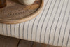 A close-up of nkuku’s Sanja Stripe Upholstered Bench in grey natural cotton, featuring dark vertical stripes, placed on light wooden flooring at the end of a bed beside a carved round wooden tray with a small vase.
