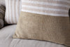 Close-up of the nkuku Samata Recycled Cushion Cover in moss, featuring horizontal cream and beige stripes, displayed on a light sofa. Another cushion in a similar design is partially visible in the background.