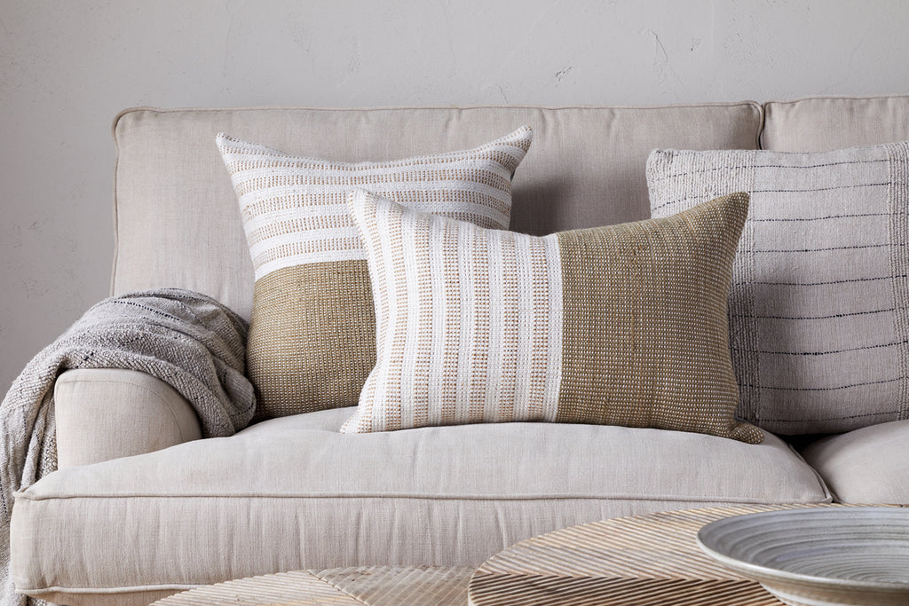 A beige sofa with two neutral pillows, including the nkuku Samata Recycled Cushion Cover in Moss, and a striped knit throw blanket on the left armrest. A round, light-colored coffee table is partially visible in front.