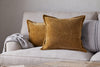Two Sela Velvet Cushion Covers in ochre by nkuku rest on a light beige sofa, accented with a gray knitted throw over one arm and a striped pillow in the background.