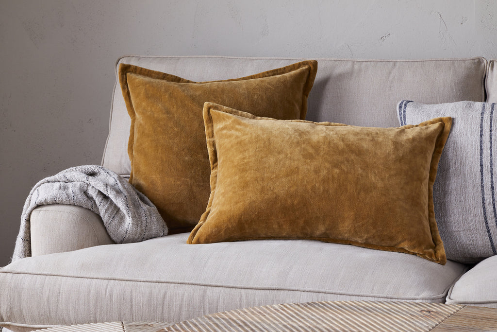 A beige sofa with two nkuku Sela Velvet Cushion Covers in Ochre, a gray knitted throw on the armrest, and a gray-striped cushion in the background add plush texture to this inviting setting.