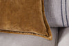 A close-up of the nkuku Sela Velvet Cushion Cover in ochre, featuring a plush texture, placed in front of a gray pillow with dark blue stripes on a light-colored fabric sofa.