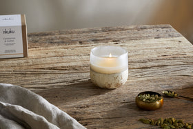 A lit nkuku Cardamom & Amber 1 Wick Scented Candle in a frosted glass holder sits on a rustic wooden table beside a brass bowl of green cardamom pods, some loose pods, a cloth, and the candle’s labeled box.