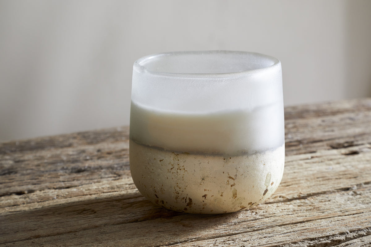 A nkuku Cardamom & Amber 1 Wick Scented Candle with a frosted glass and white-beige marbled base rests on rustic wood, filling the air with warm cardamom and amber notes against a neutral backdrop.