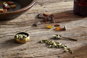 A small brass bowl, a spoon, and green cardamom pods rest on a wooden table with resin pieces and a brown bottle; pair them with the nkuku Cardamom & Amber 1 Wick Scented Candle for a warm, inviting atmosphere.