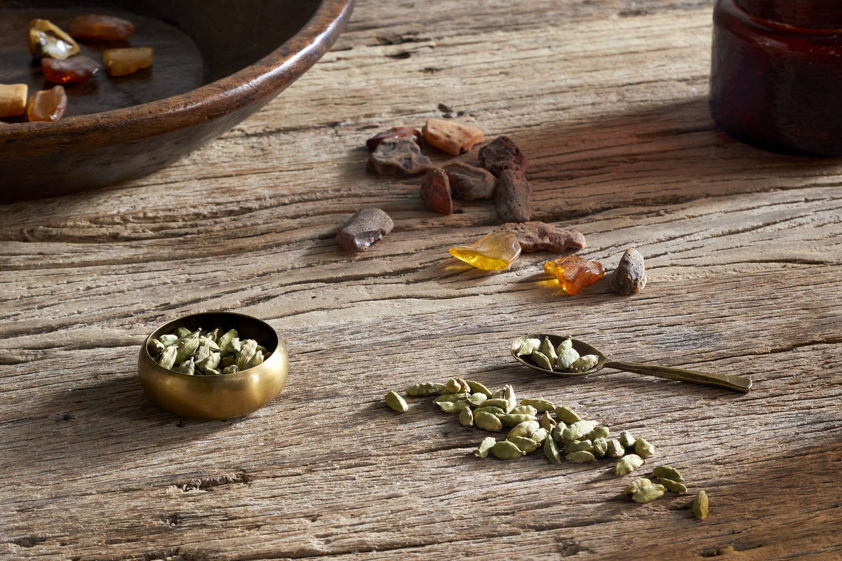 A small brass bowl, a spoon, and green cardamom pods rest on a wooden table with resin pieces and a brown bottle; pair them with the nkuku Cardamom & Amber 1 Wick Scented Candle for a warm, inviting atmosphere.