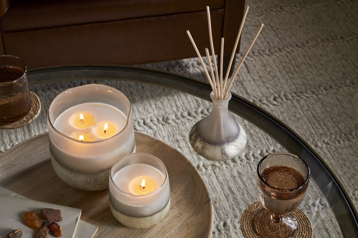 A round glass coffee table displays a nkuku Cardamom & Amber 1 Wick Scented Candle in a frosted holder, a nkuku reed diffuser, two amber glasses, and a wooden tray with stones and a folded napkin on a textured beige rug.