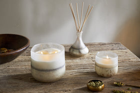 A rustic wooden table displays two nkuku Cardamom & Amber 1 Wick Scented Candles in glass holders, a small glass vase with reed diffusers, a brass bowl of cardamom pods, and a wooden bowl in the background, with sandalwood aroma lingering.