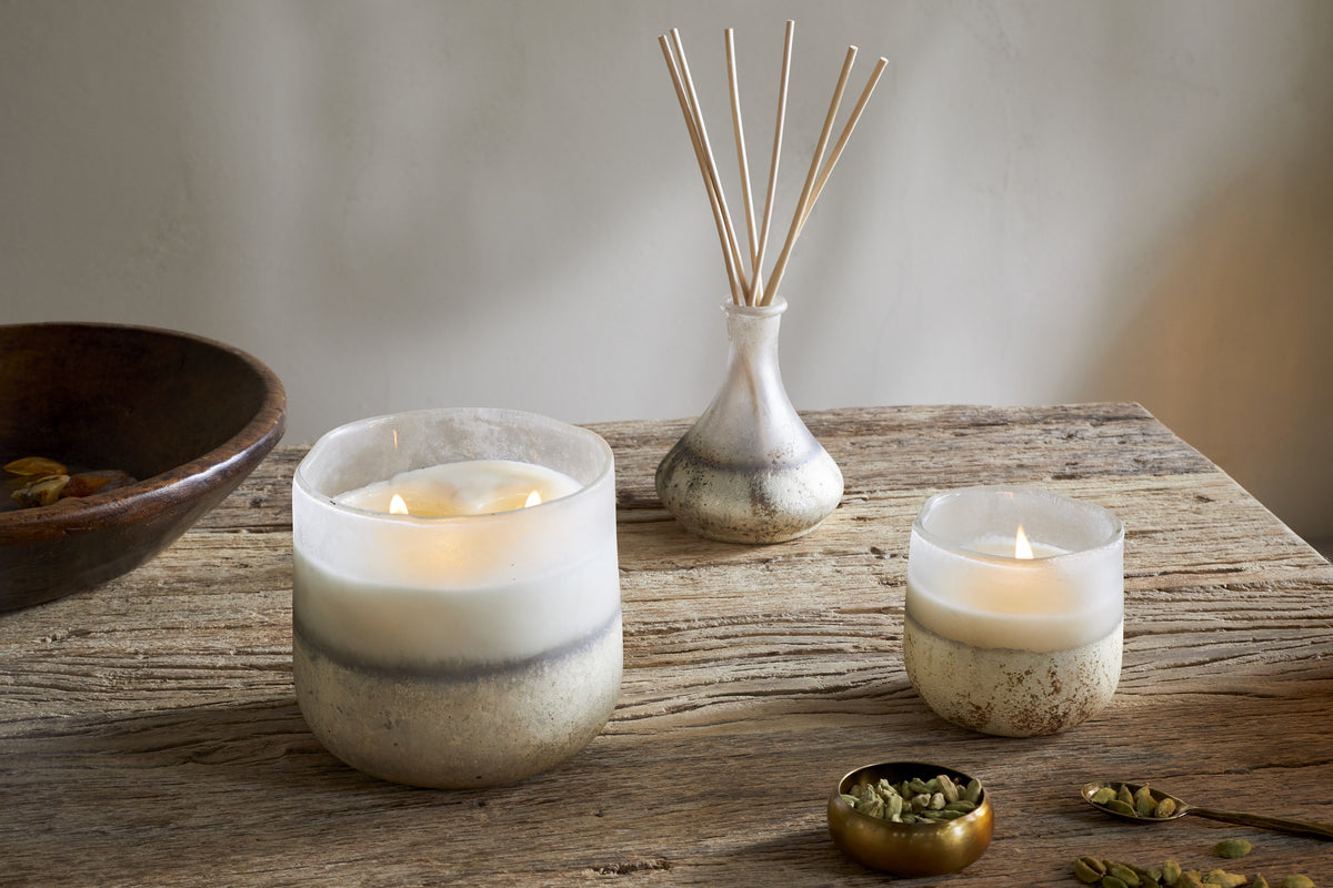 A rustic wooden table displays two nkuku Cardamom & Amber 1 Wick Scented Candles in glass holders, a small glass vase with reed diffusers, a brass bowl of cardamom pods, and a wooden bowl in the background, with sandalwood aroma lingering.