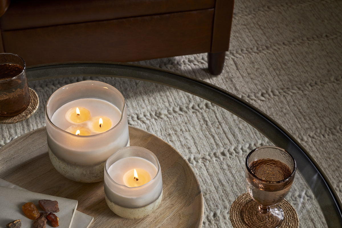 A glass coffee table displays two nkuku Cardamom & Amber 1 Wick Scented Candles in glass jars on a wooden tray, with a drink on a coaster and a folded napkin with stones, next to a beige textured rug and brown armchair.