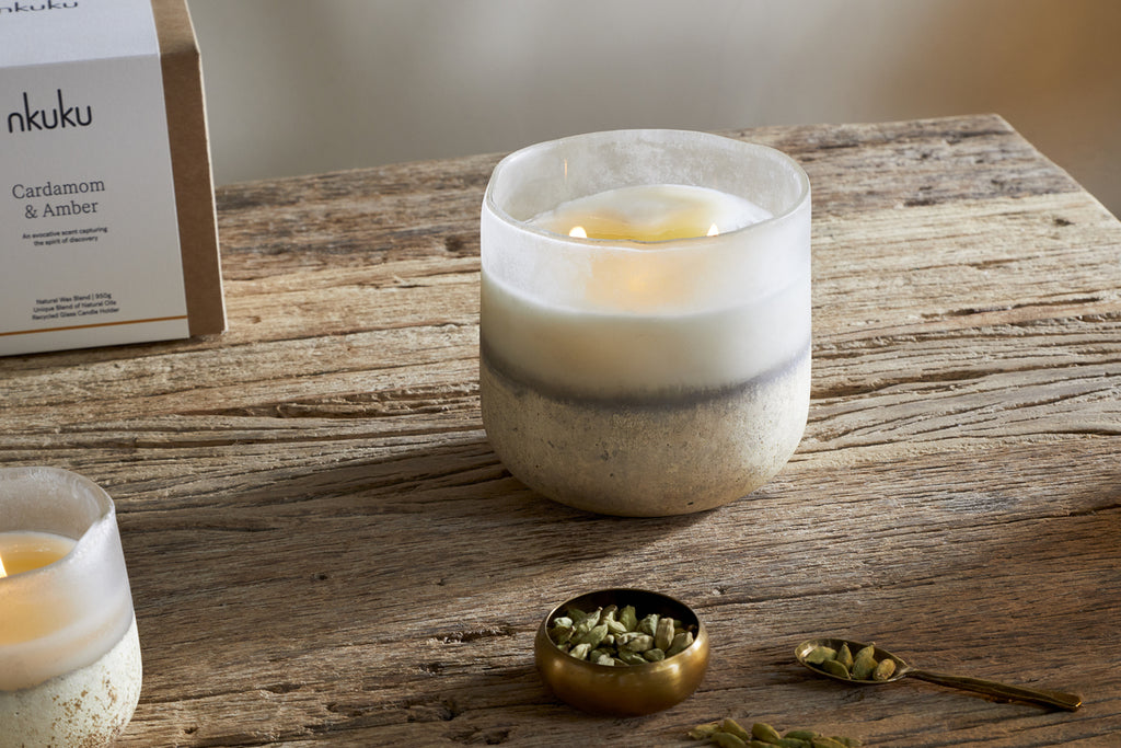 A nkuku Cardamom & Amber 3 Wick Scented Candle in frosted glass rests on a rustic wooden table by a brass bowl of cardamom pods, evoking sandalwood notes, with its open box visible in the background.