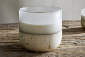 A frosted glass nkuku Cardamom & Amber 3 Wick Scented Candle with a white and beige gradient and rustic texture sits on a weathered wooden table.