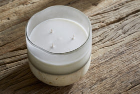The nkuku Cardamom & Amber 3 Wick Scented Candle, in a frosted glass container, rests on a rustic wooden surface and fills the air with its warm, inviting fragrance.