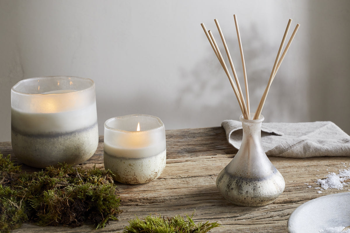 Two lit nkuku Sea Salt & Moss 1 Wick Scented Candles in ceramic holders rest on a rustic wooden table, accented with green moss and a folded beige cloth, creating a cozy ambiance filled with bespoke fragrance.