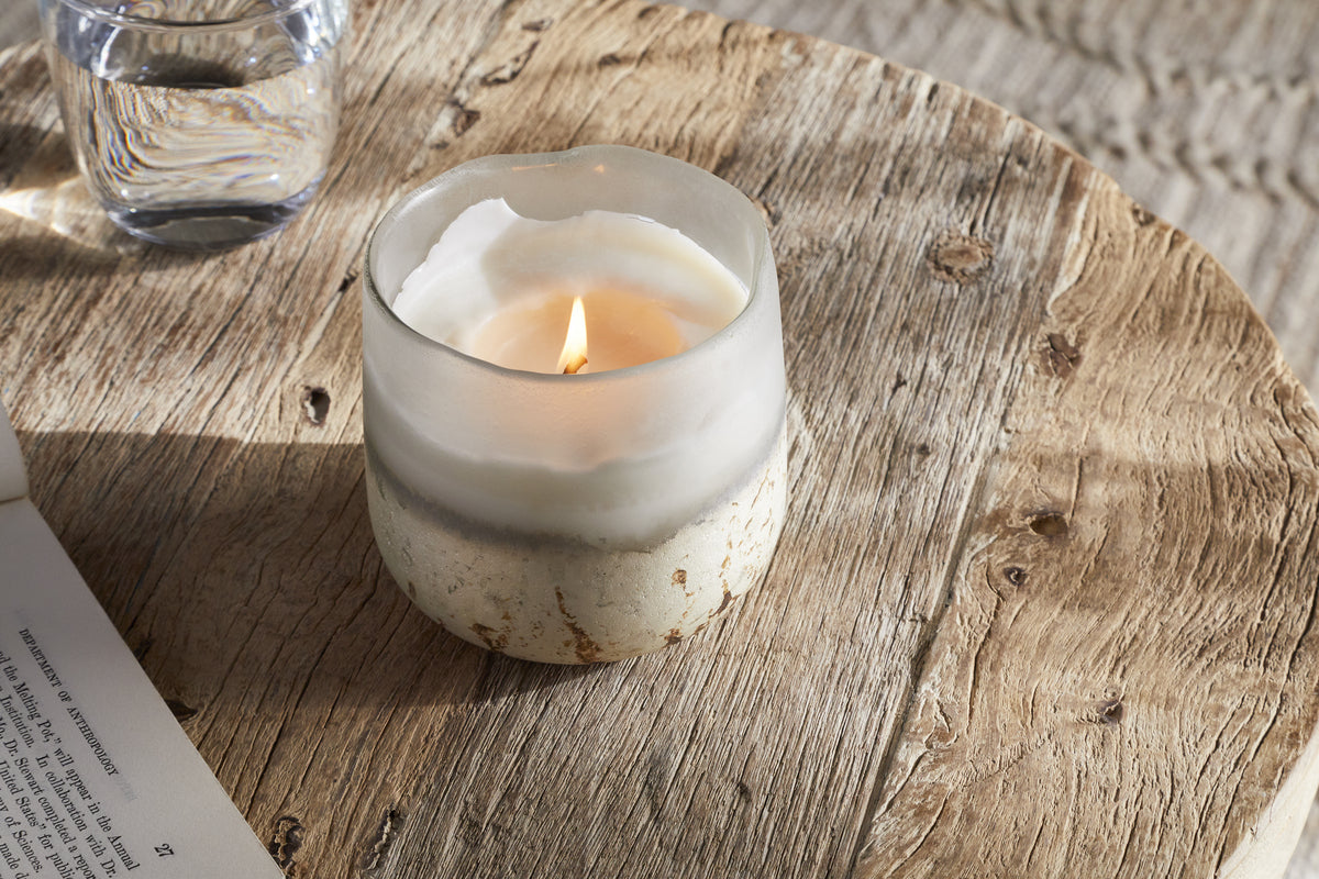 The nkuku Sea Salt & Moss 1 Wick Scented Candle in a frosted glass holder sits on a rustic round wooden table, beside a glass of water and an open book, as sunlight casts soft shadows over the textured wood surface.
