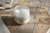 A nkuku Sea Salt & Moss 1 Wick Scented Candle in a frosted glass holder rests on a rustic wooden table beside a glass of water and an open book, as sunlight casts gentle shadows over the scene.