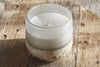 The nkuku Sea Salt & Moss 1 Wick Scented Candle, in a frosted glass holder with a textured base, rests on rustic wood and fills the air with its unique fragrance.