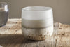 The nkuku Sea Salt & Moss 1 Wick Scented Candle in a frosted glass holder with a rustic, textured base sits on a wooden table, softly diffusing its bespoke fragrance in natural light.