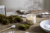 A nkuku Sea Salt & Moss 1 Wick Scented Candle in a frosted holder rests on a rustic wooden table with moss and a beige cloth, adding bespoke fragrance for a natural, calming atmosphere.
