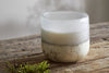 A nkuku Sea Salt & Moss 1 Wick Scented Candle with a cream and gray marbled base rests on a rustic wooden surface, its bespoke fragrance filling the air, with green moss in the lower left corner.