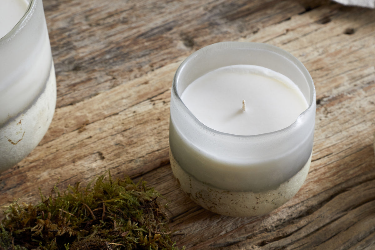 The nkuku Sea Salt & Moss 1 Wick Scented Candle in a frosted glass container rests on rustic wood near green moss, infusing the air with its bespoke fragrance.