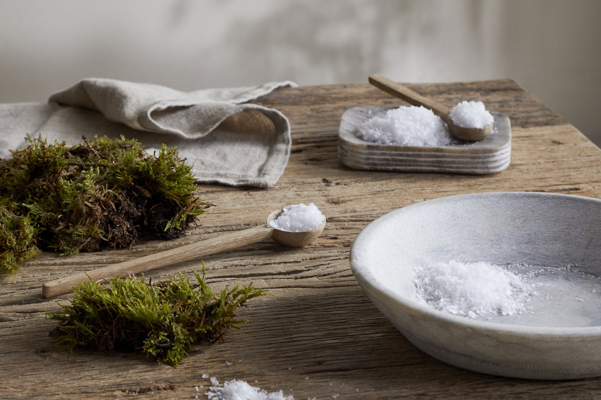 A rustic wooden table displays a white napkin, green moss, a bowl of coarse salt, and small spoons—perfect for highlighting the nkuku Sea Salt & Moss 3 Wick Scented Candle, crafted in the UK.