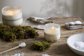 The nkuku Sea Salt & Moss 3 Wick Scented Candle in a glass holder fills a rustic wooden table with tranquil fragrance, surrounded by green moss, linen napkin, and wooden salt dishes—adding natural charm to any UK home.
