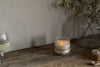 The nkuku Sea Salt & Moss 3 Wick Scented Candle glows in its glass holder on a rustic wooden table beside a glass of white wine, green stems in a clear vase, and a small wooden bowl—a cozy UK-inspired scene.