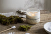 The nkuku Sea Salt & Moss 3 Wick Scented Candle in a frosted glass holder sits on a rustic wooden table with moss, a small wooden spoon of salt, a folded cloth, and a white ceramic bowl filled with salt.