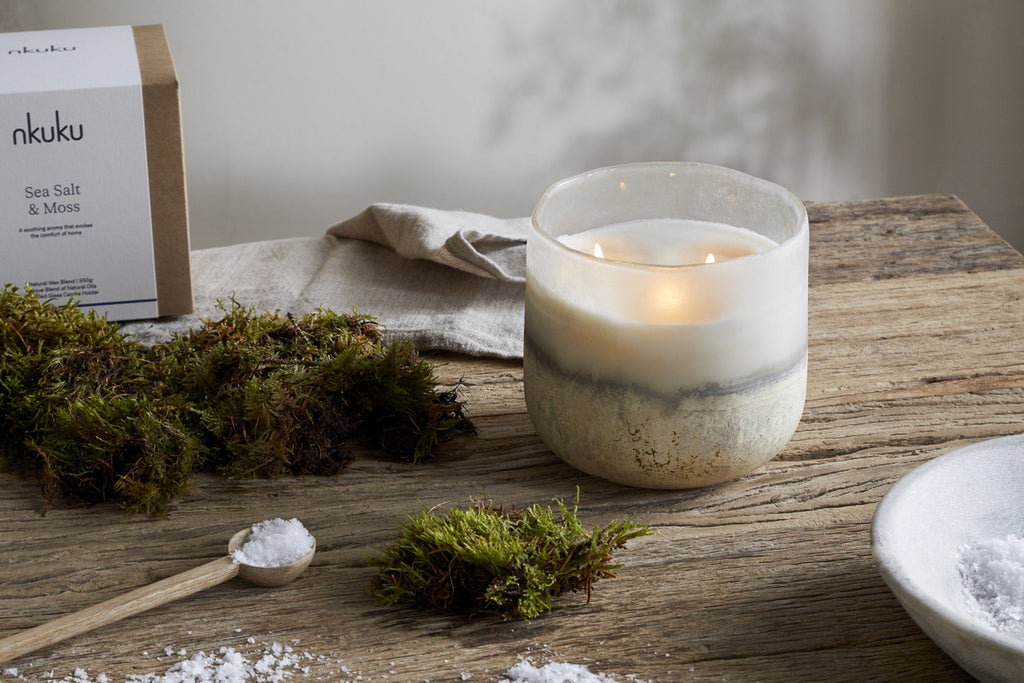 The nkuku Sea Salt & Moss 3 Wick Scented Candle, in a white and gray marbled holder, rests on a rustic wooden table beside green moss, sea salt, and its box—evoking a bespoke UK-inspired fragrance.