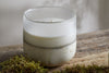 The nkuku Sea Salt & Moss 3 Wick Scented Candle, housed in a frosted glass container with a rustic textured base, rests on a weathered wooden surface beside green moss.