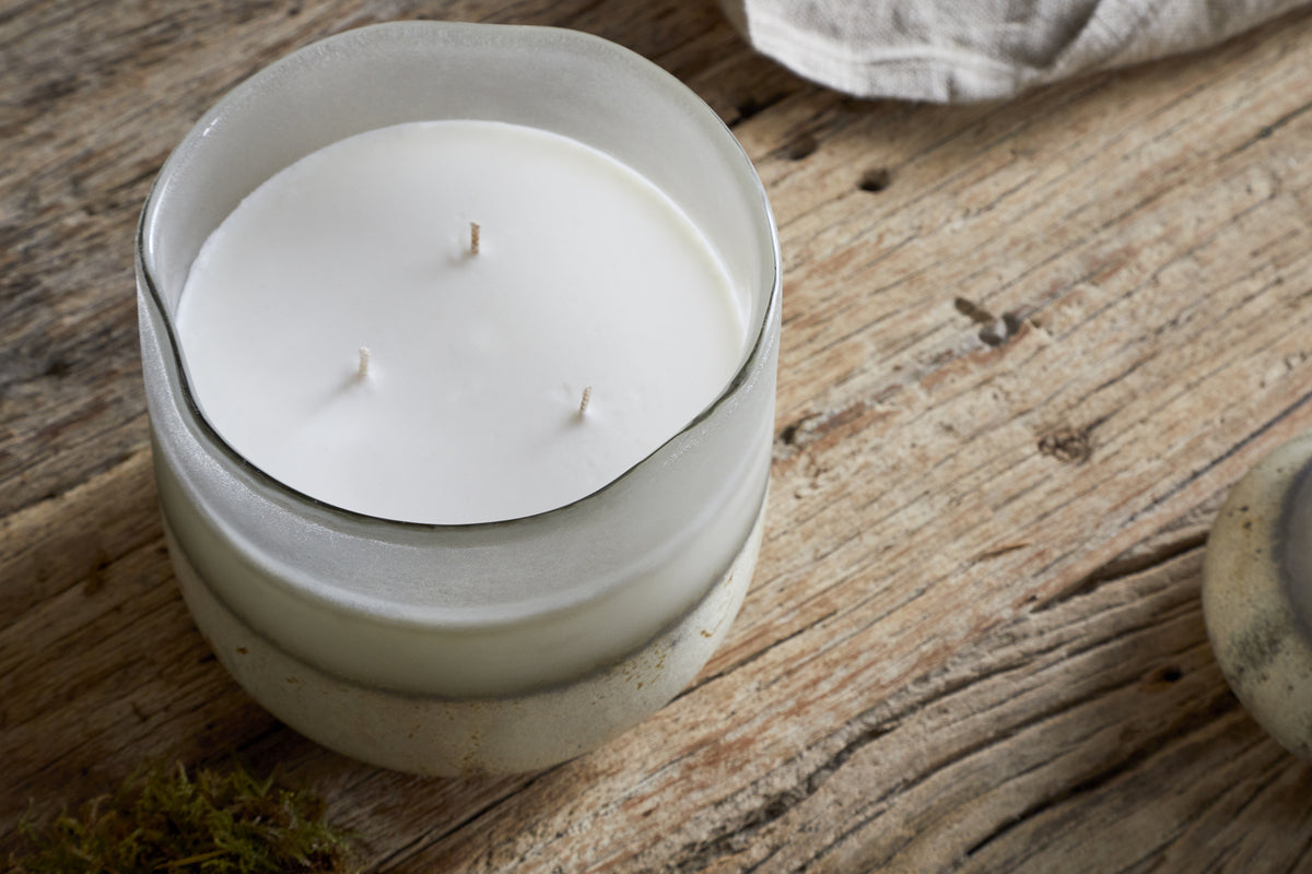 The nkuku Sea Salt & Moss 3 Wick Scented Candle, in a frosted glass jar, rests on a rustic wooden surface with a hint of light fabric in the background.
