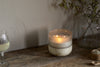 The nkuku Sea Salt & Moss 3 Wick Scented Candle in a ceramic holder sits on a rustic wooden table beside a glass of white wine, a clear jar with dried branches, and a small wooden bowl—ideal for cosy UK evenings.
