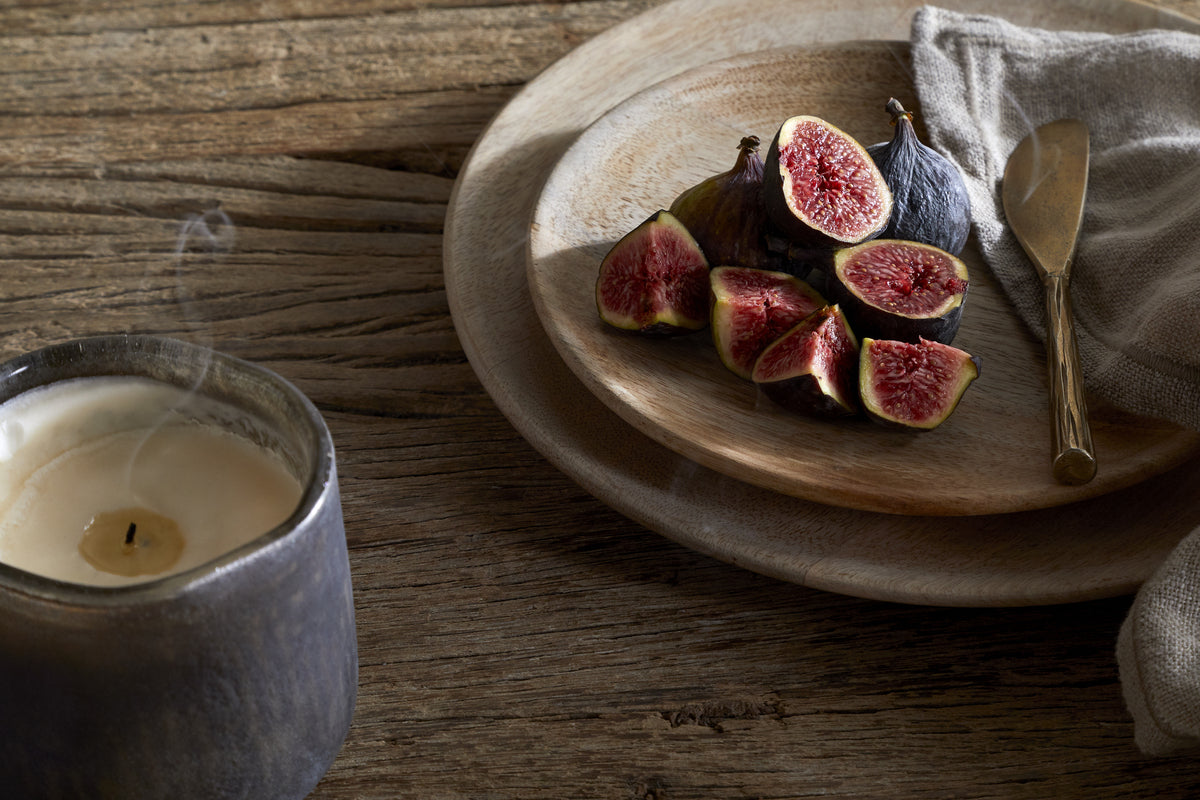 A sliced fig on a wooden plate with a gold knife and beige napkin sits beside a lit candle in a ceramic holder and the nkuku Cedarwood & Fig Reed Diffuser, all arranged on a rustic wooden table.