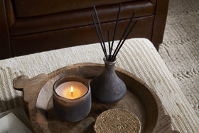 A lit candle, a small woven coaster, and the nkuku Cedarwood & Fig Reed Diffuser sit on a round wooden tray atop a textured beige ottoman, with a brown leather sofa in the background.