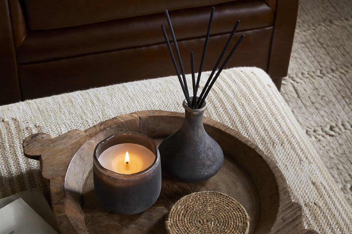 The nkuku Cedarwood & Fig 1 Wick Scented Candle, a small vase with black diffuser sticks, and a woven coaster rest on a round wooden tray over textured beige fabric, with a brown leather chair in the background.