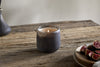 A lit nkuku Cedarwood & Fig 1 Wick Scented Candle in a rustic glass holder sits on a weathered wooden table, infusing the air with cedarwood and fig notes beside a ceramic plate of sliced figs.