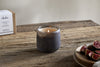 A lit nkuku Cedarwood & Fig 1 Wick Scented Candle in a dark glass jar sits on a rustic table, with its box to the left and sliced figs to the right, highlighting its honeyed fig and cedarwood fragrance.