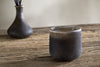 The Cedarwood & Fig 1 Wick Scented Candle by nkuku, a small dark ceramic piece, sits on a rustic wooden table with a blurred vase and black reeds behind it, creating a minimalist and earthy vibe.