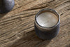 The nkuku Cedarwood & Fig 1 Wick Scented Candle, in a dark rustic ceramic holder, sits on a textured wooden surface. The white candle is unused with its wick still intact.