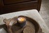 The nkuku Cedarwood & Fig 1 Wick Scented Candle in a dark glass holder rests on a round wooden tray with a woven coaster atop a textured beige ottoman, with a brown leather sofa and woven rug in the background.
