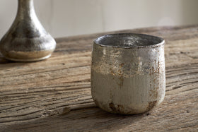 A Juniper & Fir 1 Wick Scented Candle by nkuku, in a weathered metallic holder with a textured finish, sits on a rustic wooden table. A blurred silver vase appears in the background.