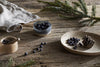 A cozy scene with scattered juniper berries, a small wooden bowl and spoon, a ceramic bowl of berries, pine branches, a folded cloth, and the nkuku Juniper & Fir 1 Wick Scented Candle in a reusable vessel.