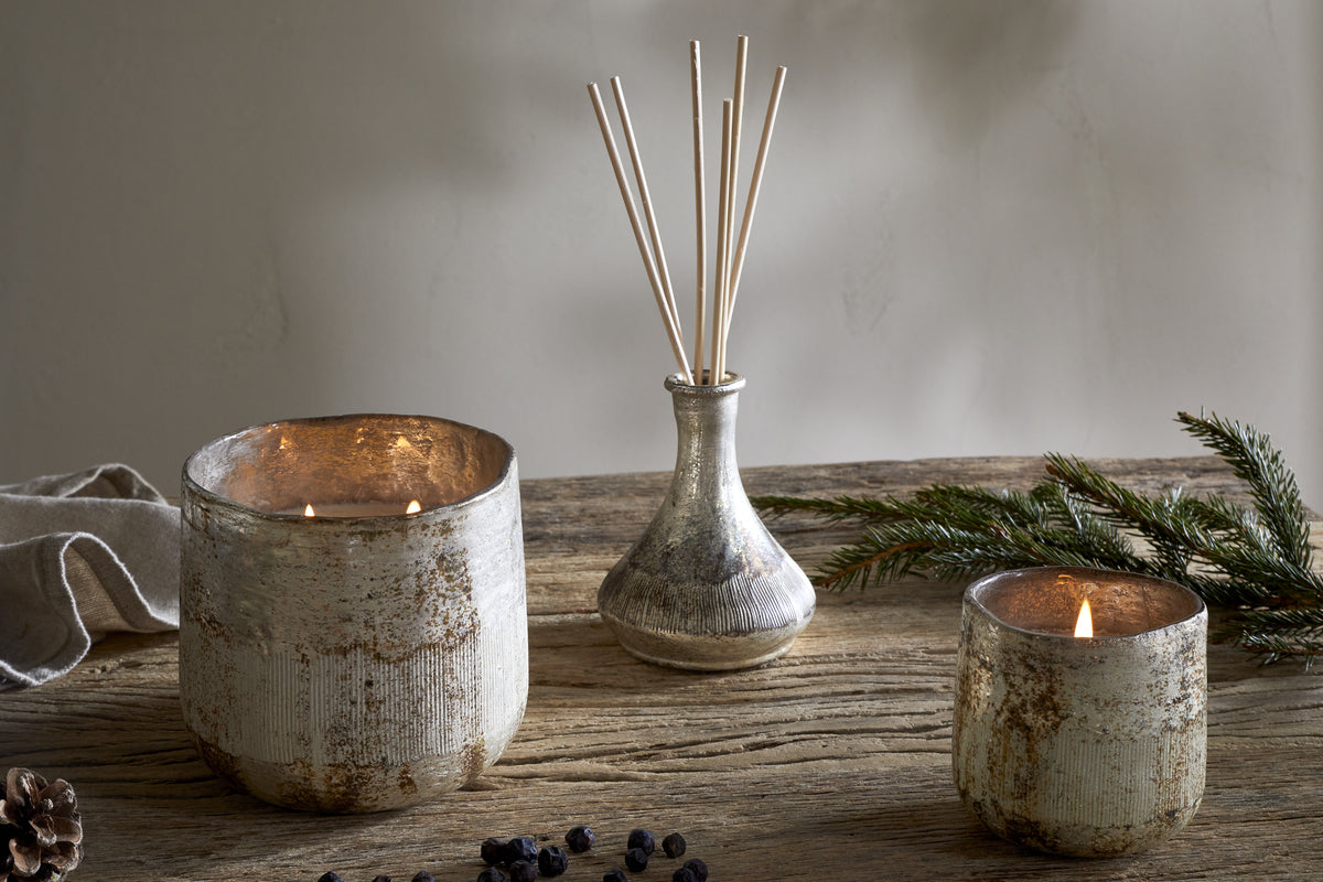 A rustic wooden table features the nkuku Juniper & Fir 1 Wick Scented Candle in a textured, reusable vessel, paired with two lit candles, a glass vase of reed diffuser sticks, pine branches, pinecones, and berries.