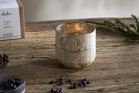 The nkuku Juniper & Fir 3 Wick Scented Candle in a rustic glass holder sits on a wooden table with juniper berries, a pinecone, and fir branch—a cozy display that captures the essence of winter.