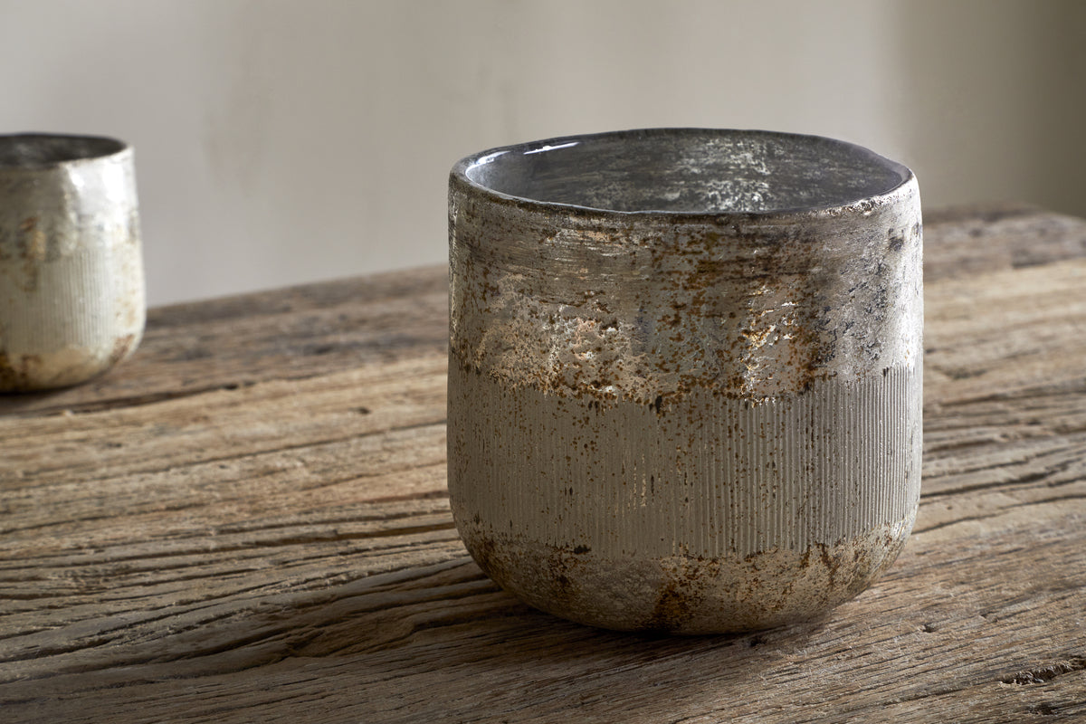 The nkuku Juniper & Fir 3 Wick Scented Candle rests on a rustic wooden table, its earthy ceramic vessel evoking the calm of winter; another similar candle is blurred behind, suggesting a serene, minimalist space.
