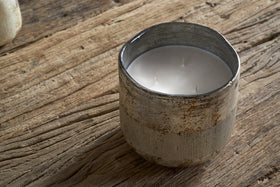 The nkuku Juniper & Fir 3 Wick Scented Candle, with a bespoke fragrance, sits in a rustic textured ceramic holder on a weathered wooden surface.