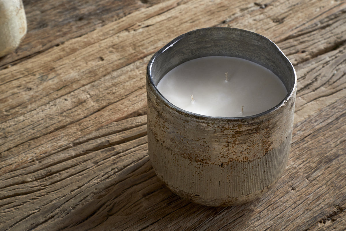 The nkuku Juniper & Fir 3 Wick Scented Candle, with a bespoke fragrance, sits in a rustic textured ceramic holder on a weathered wooden surface.